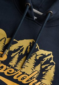 Marineblå hoodie med et tekstureret gult bjerg- og træmotiv, der har mørke snore med metalspidser indgraveret med "Superdry."