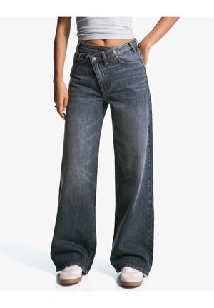 Personne portant un jean en denim sombre à jambes larges avec une ceinture asymétrique qui se chevauche et des baskets blanches, debout sur fond blanc.