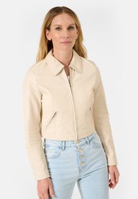 Getailleerde beige leren jas met een kraag en ritssluiting, voorzien van zijzakken met rits en gestructureerde stof. Draagt over lichtblauwe high-waisted jeans.
