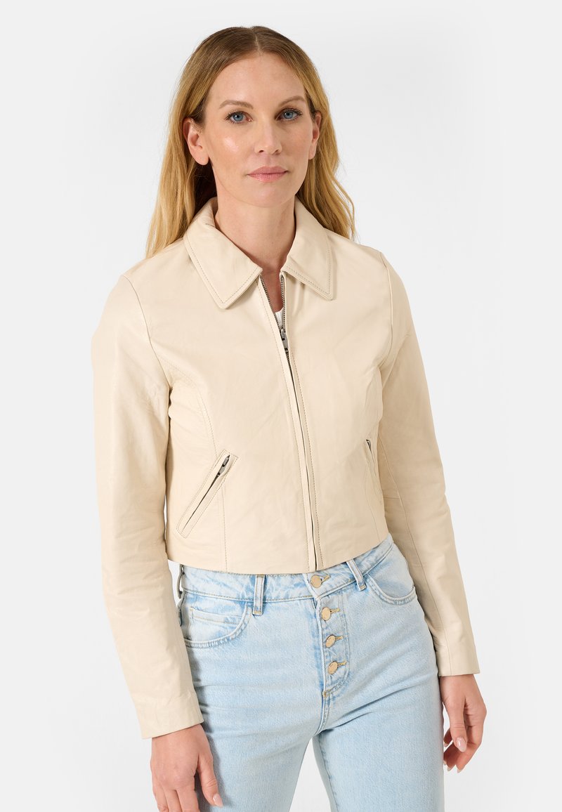 Getailleerde beige leren jas met een kraag en ritssluiting, voorzien van zijzakken met rits en gestructureerde stof. Draagt over lichtblauwe high-waisted jeans.
