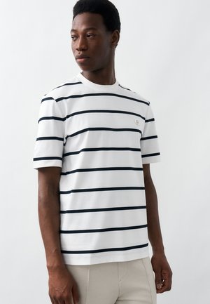 Άνδρας που φοράει λευκό κοντομάνικο μπλουζάκι με οριζόντιες μαύρες ρίγες, κοιτάζοντας αριστερά πάνω σε απλό λευκό φόντο.