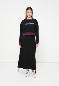 Svart avkortet sweatshirt med lilla detaljer og "SANTA CRUZ" logo, kombinert med et langt svart skjørt og hvite sko med stropper.