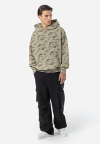 Haut à capuche camouflé kaki avec une poche avant et un cordon de serrage. Associé à un pantalon cargo noir ample et des baskets blanches.