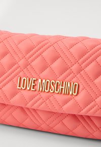 Pletená korálová kožená kabelka přes rameno s výrazným zlatým logem "LOVE MOSCHINO" umístěným na přední straně. Texturovaný vzor přidává hloubku.