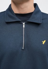 Tmavomodrá mikina na zips z mäkkej látky, s strieborným zipsom a malým žltým logom vtáčika na ľavej strane hrudníka.