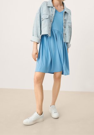 Mujer que lleva un vestido azul hasta la rodilla con cuello en V, chaqueta vaquera corta de lavado claro y zapatillas blancas, de pie sobre un fondo neutro.