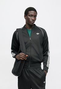 Juodas sportinis švarkelis su baltomis juostelėmis, užtrauktuku užsegamas ir logotipu. Derinamas su atitinkančiomis kelnėmis ir juoda petnešine rankine. Po apačia žalia marškinėliai.