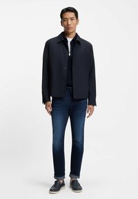 Man standing wearing a dark navy jacket over a zipped sweater, dark blue rolled-up jeans, and navy slip-on shoes against a plain background.