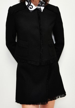 Femme portant un tailleur noir texturé en deux pièces avec des bordures à franges et une chemise blanche avec un motif floral noir sur le col.