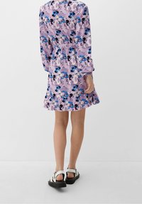 Robe florale violette à manches longues, taille cintrée, ourlet froncé et accents noirs. Portée avec des sandales blanches et noires, mettant en avant la vue arrière.