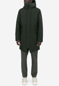 Mörkgrön huva med dragsko, framzip, två sidofickor och rak skärning. Tyget är slätt med en matt yta.