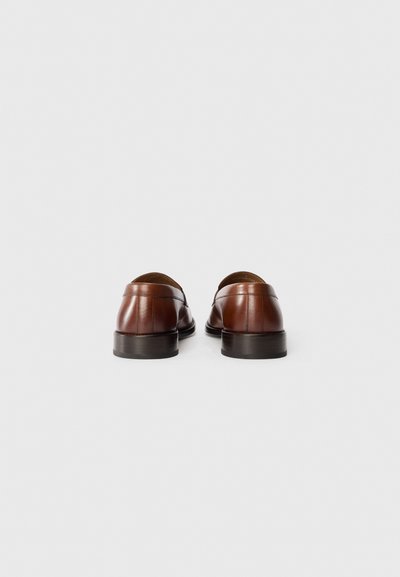 Mocasines de cuero marrón con un acabado liso, tacones bajos apilados y detalles de costura sutiles. El diseño incluye una punta redonda y un estilo de calzado fácil de poner.