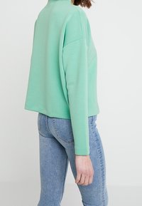 Mintgrön sweatshirt med hög krage, oversized passform och långa ärmar, kombinerad med ljusblå jeans. Mjuk textur, avslappnad design.