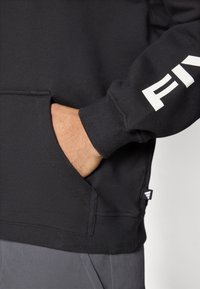 Sweatshirt preta com bolso canguru, punhos canelados e um detalhe de logótipo branco na manga. O tecido aparenta ser macio com uma textura suave.