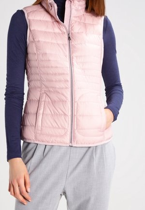 Femme portant une veste matelassée rose clair sur une chemise bleu marine à manches longues avec un pantalon gris clair, une main dans la poche de la veste sur un fond uni.