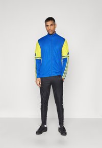 Blauwe en gele sportjas met rits, lange mouwen en zwarte trainingsbroek. Bevat Adidas-strepen en logo-accenten.