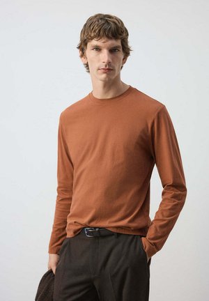 Jeune homme aux cheveux bruns courts portant une chemise à manches longues couleur rouille et un pantalon foncé sur un fond gris uni.