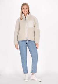 Giacca in pile di un beige chiaro con zip frontale, tessuto textured e tasca sul petto. Indossata con jeans blu e sneakers bianche alte.