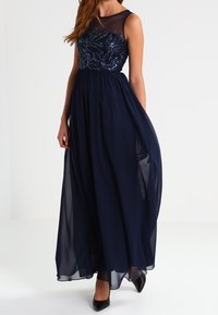 Mörkblå aftonklänning med en figursydd paljettliv, genomskinlig halsringning och flytande chiffongkjol. Svarta klackar fulländar looken.