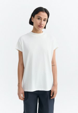 Woman with short dark hair wearing a plain white short-sleeve shirt and dark pants, standing against a white background.