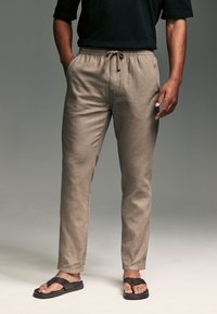Pantaloni beige, leggeri, a gamba affusolata con vita elastica e coulisse regolabile. Indossati con ciabatte nere.