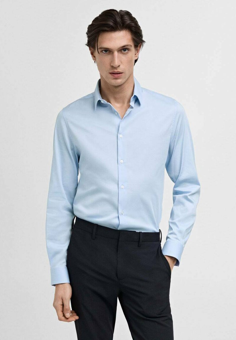 Chemise bleu clair à manches longues, devant boutonné et col bien net, assortie avec un pantalon sombre. Texture lisse et coupe ajustée.