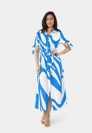 Donna che indossa un vestito midi con bottoni, maniche corte e motivo astratto blu e bianco, con cintura in vita, in piedi davanti a uno sfondo semplice.