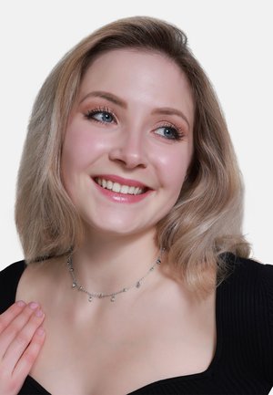 Srebrny naszyjnik łańcuszkowy z wieloma małymi zawieszkami, noszony na ciemnej bluzce. Modelka ma jasnobrązowe włosy i uśmiechniętą minę.