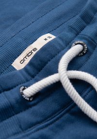 Blåt stofbånd med hvid snor, der er træde gennem sorte metaløjer, og et hvidt mærke med "ombre" mærket og størrelse M.