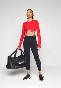 Rode crop top met lange mouwen en een witte "NIKE PRO" band, zwarte legging met witte zijstrepen, en zwarte duffeltas met wit logo. Zwarte sneakers.