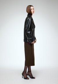 Melna ādas motociklista jaka ar sudraba rāvējslēdzējiem, brūna ceļgalu garuma svārki, melni strapa papēži un caurspīdīgas melnas zeķubikses. Skats no sāniem.