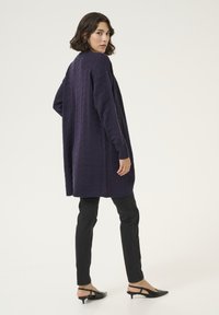 Langer, übergroßer Cardigan in Dunkelpurpur mit einem strukturierten Zickzackmuster. Kombiniert mit schwarzen Slim-Fit-Hosen und schwarzen Slingback-Absätzen.