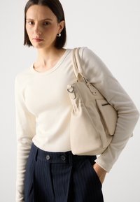Beige skinnveske med polert maskinvare, parret med en lys kremfarget langermet skjorte og marineblå stripete bukser.