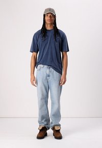 Temno modra majica s kratkimi rokavi, svetlo modri džinsi in rjavi škornji. Oseba nosi tudi sivo kape in ima dolge, pletene lase.