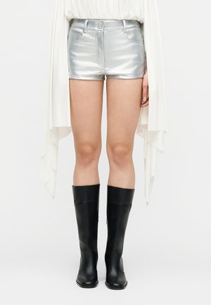 Person iført metallic sølvshorts, hvid langærmet top med forlængede ærmer og sorte knæhøje støvler, stående oprejst.