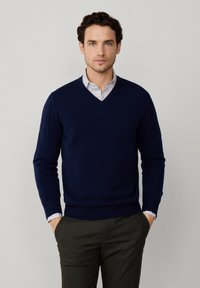 Granatowy sweter w serek z miękkiej dzianiny, z długimi rękawami i ściągaczami, noszony na jasnoniebieskiej koszuli w paski.