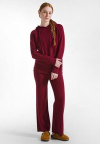 Ensemble de loungewear bourgogne composé d'un hoodie à manches longues avec une poche avant et d'un pantalon ample, associé à des pantoufles intérieures marron.