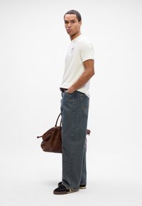 Creme bomulds t-shirt med logo, løstsiddende blå denim jeans, sneakers i sort og tan, og en brun lædertaske. Simpelt design og minimale detaljer.