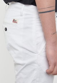 Pantaloni bianchi in cotone con una tasca posteriore dotata di un bottone nero e di un patch con logo. Il tessuto appare liscio e leggermente stropicciato.