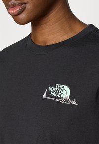 Μαύρο βαμβακερό T-shirt με ανοιχτό μπλε γραφικό λογότυπο της εταιρείας The North Face, που περιλαμβάνει σχέδιο σκηνής και δέντρου, στο αριστερό μέρος του στήθους.