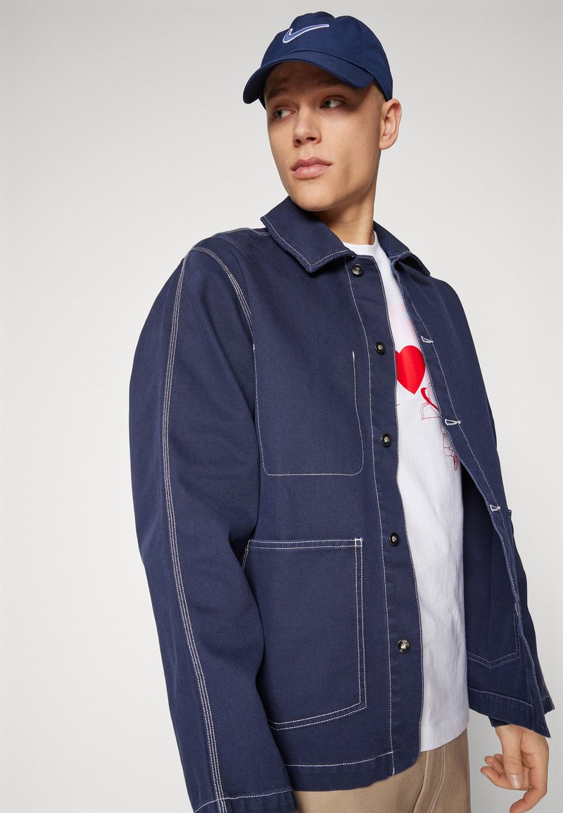 Chaqueta de mezclilla azul marino con costuras blancas, que cuenta con un cuello, botones frontales y dos grandes bolsillos de parche. Usada sobre una camiseta blanca.