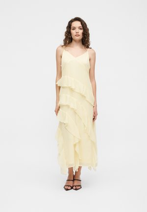 Femme aux cheveux bouclés portant une robe longue sans manches jaune pâle avec des volants superposés et des talons noirs à brides, debout devant un fond clair.