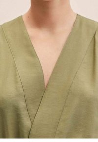 Top cruzado verde oliva con un escote en V profundo, hecho de tela suave. Presenta detalles sutiles de costura a lo largo de las costuras.