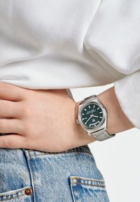 Montre en acier inoxydable argenté avec un boîtier octagonal, un cadran vert, des index blancs et un affichage de la date, portée au poignet sur un jean en denim bleu clair.