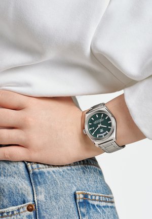 Montre en acier inoxydable argenté avec un boîtier octagonal, un cadran vert, des index blancs et un affichage de la date, portée au poignet sur un jean en denim bleu clair.