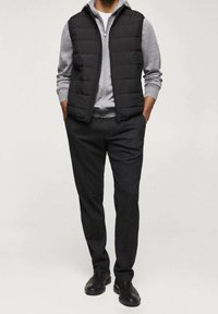 Schwarze Puffer-Weste über einem grauen Hoodie mit Reißverschluss, kombiniert mit dunkelgrauen Hosen. Glatte Texturen und minimalistisches Design, ergänzt durch schwarze Schuhe.