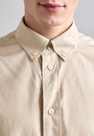 Beige skjorte med knapper og standard krave, der har en glat overflade, en stolpe med tre knapper og syede detaljer.