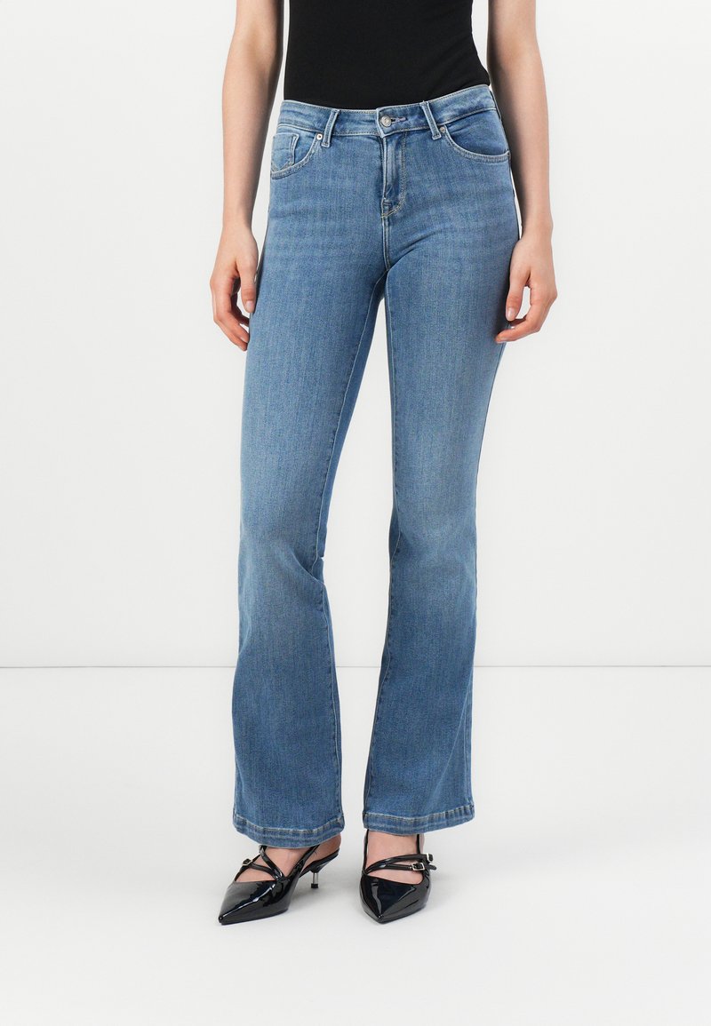 Kvinna i ljusblå bootcut-jeans och svarta klackskor med spetsig tå och remmar, stående mot en enfärgad vit bakgrund.