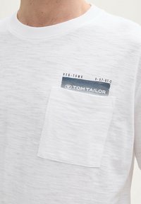 Hvid kortærmet t-shirt med en tekstureret overflade, der har en lomme med en blå label, rund halsudskæring og afslappet pasform.