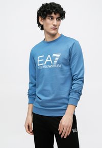 Μπλε φούτερ με μακριά μανίκια, στρογγυλή λαιμόκοψη και λευκό λογότυπο "EA7 EMPORIO ARMANI". Φοριέται με μαύρο παντελόνι. Υλικό από μείγμα βαμβακιού.